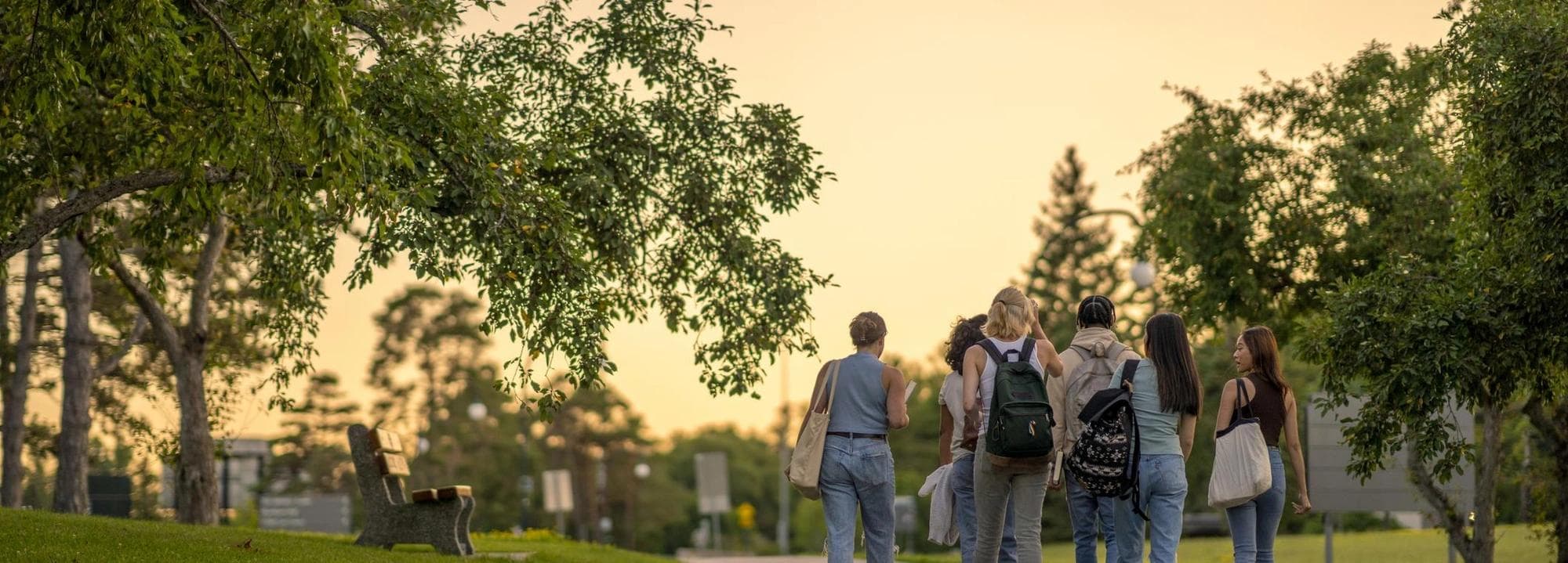 A group of University students are seen from behind walking outside on campus as they make their way to class. They are dressed casually and have backpacks on as they talk together and make their way to school for the day.