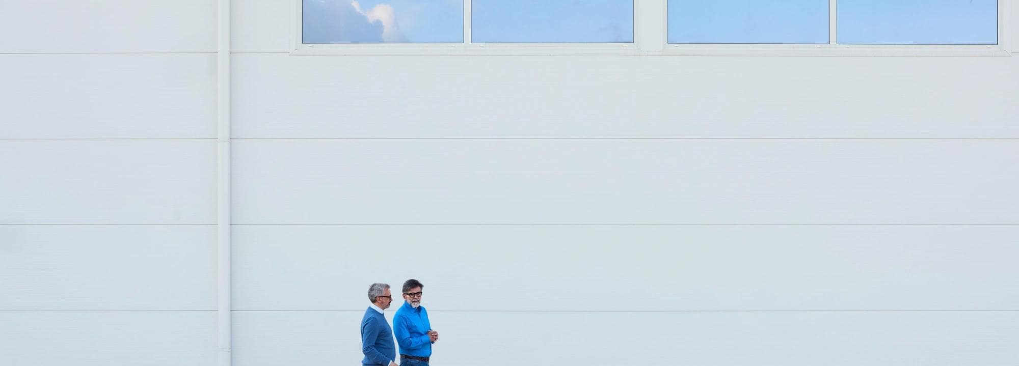 Two individuals walk near a modern white building with clear architectural lines.